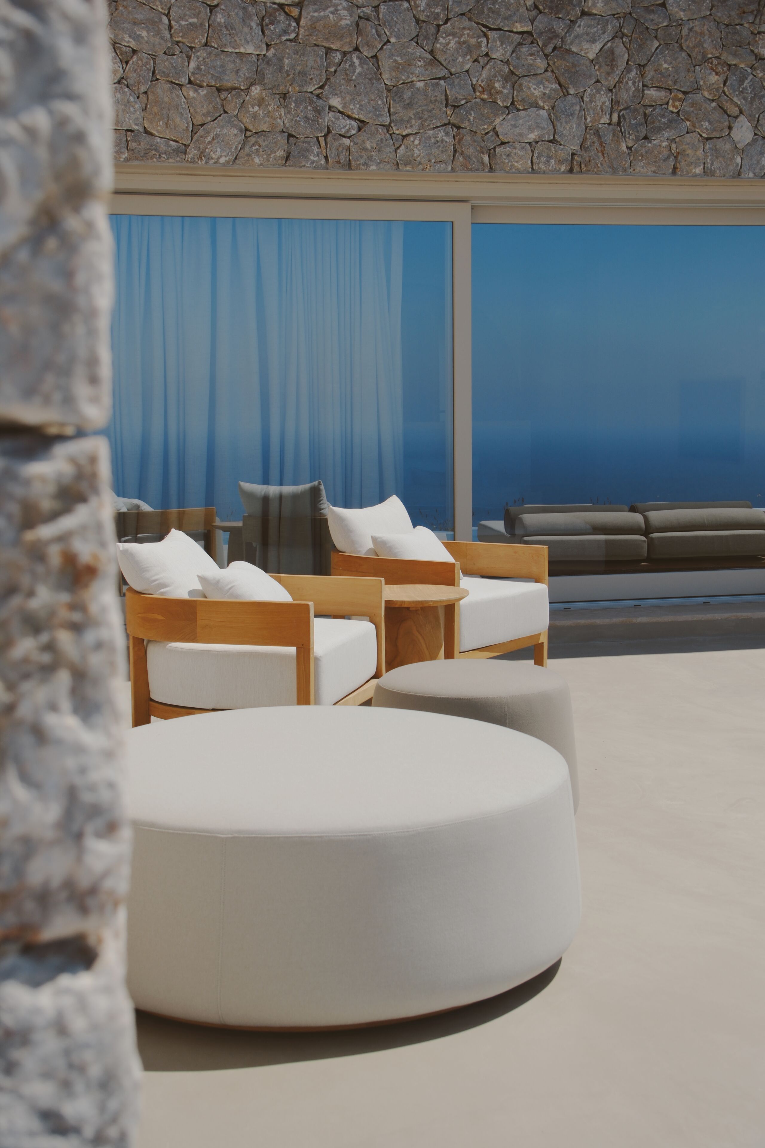 A modern outdoor seating area with a stone wall backdrop, a large window with blue curtains, and a view of the ocean.