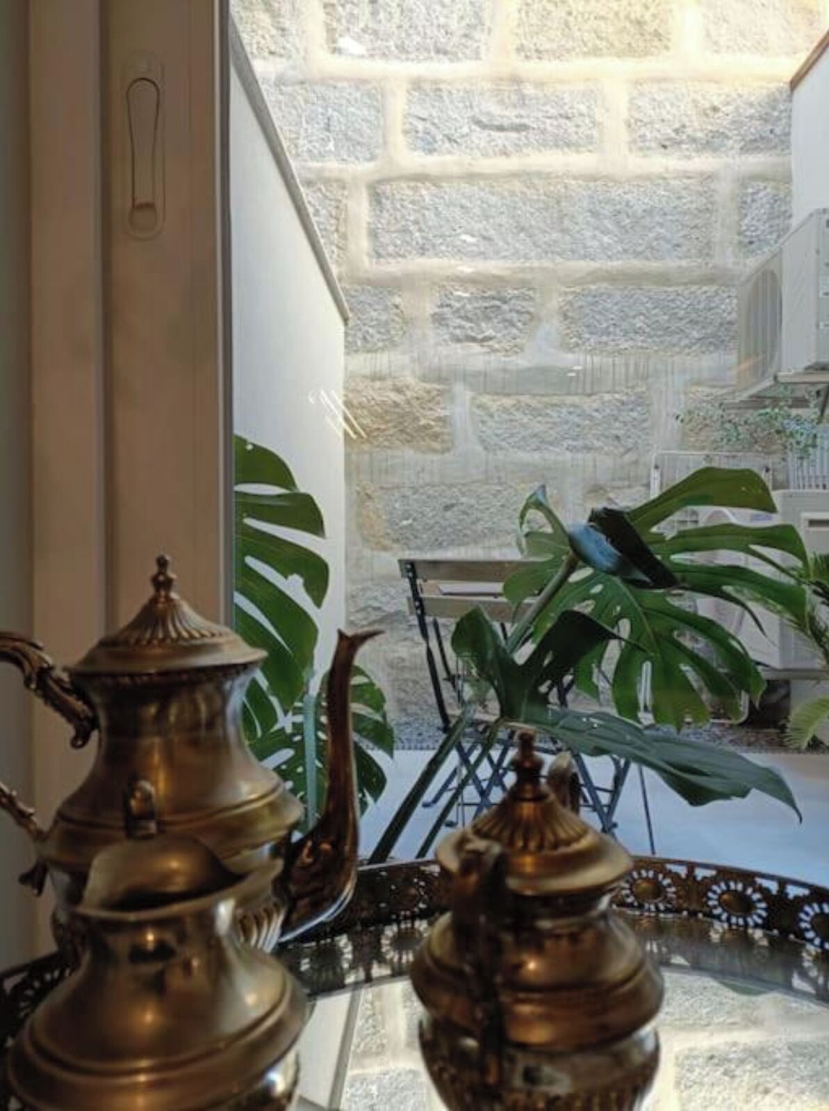 A brass teapot and a decorative item on a table, with a view of a stone wall and green plants outside.