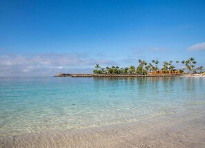 A clear turquoise sea with a sandy beach and a small island with palm trees and buildings. - Duplex in Maspalomas Near Meloneras Beach (San Bartolomé de Tirajana)