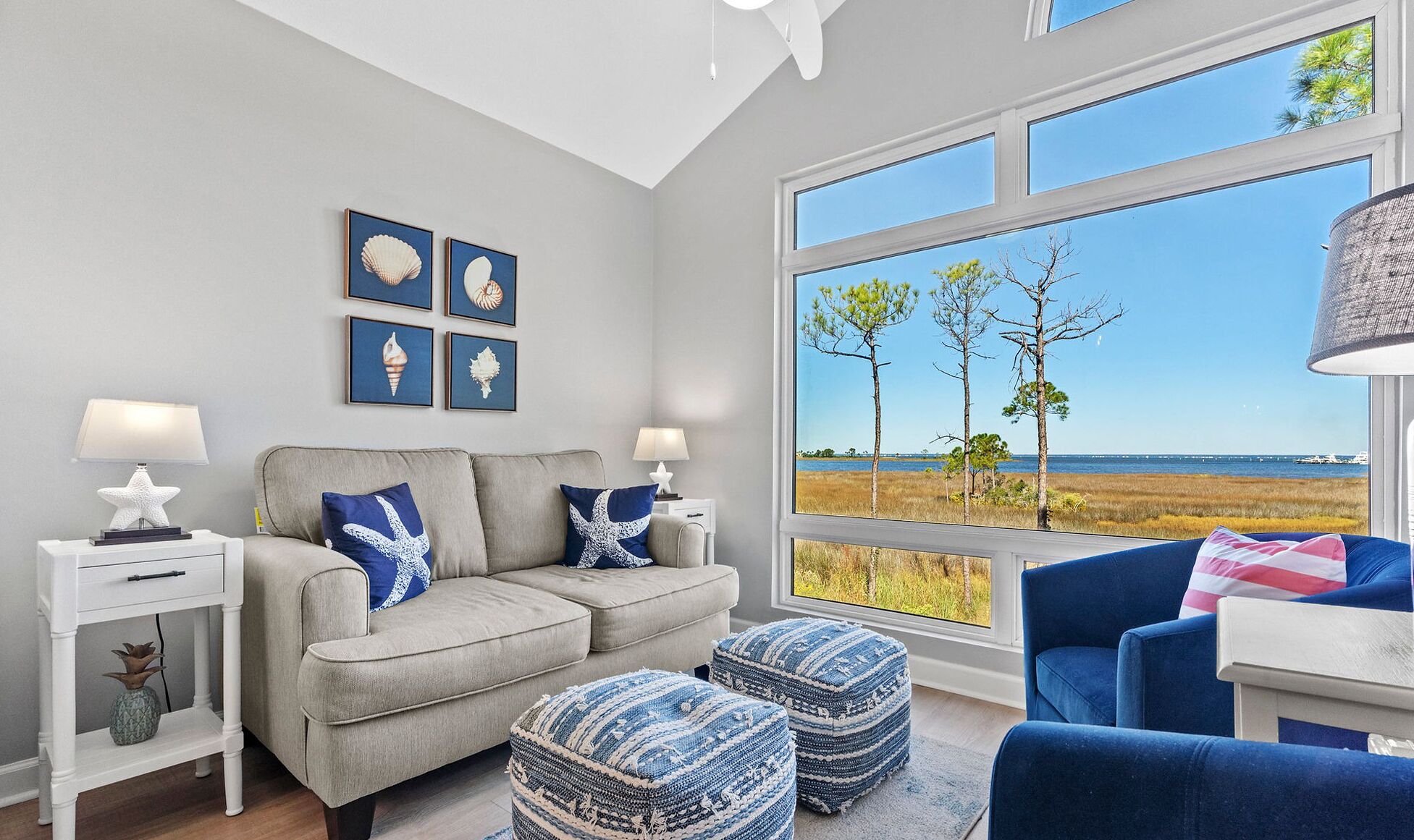 Een woonkamer met een grijze bank, blauw-witte kussens, een witte bijzettafel en een groot raam met uitzicht op bomen en water.