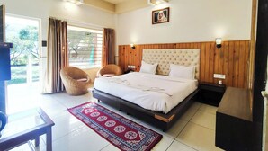 A bedroom with a large bed, a ceiling fan, a window with curtains, a wicker chair, and a red patterned rug. - MVT Resort & Restaurant (Rishikesh)