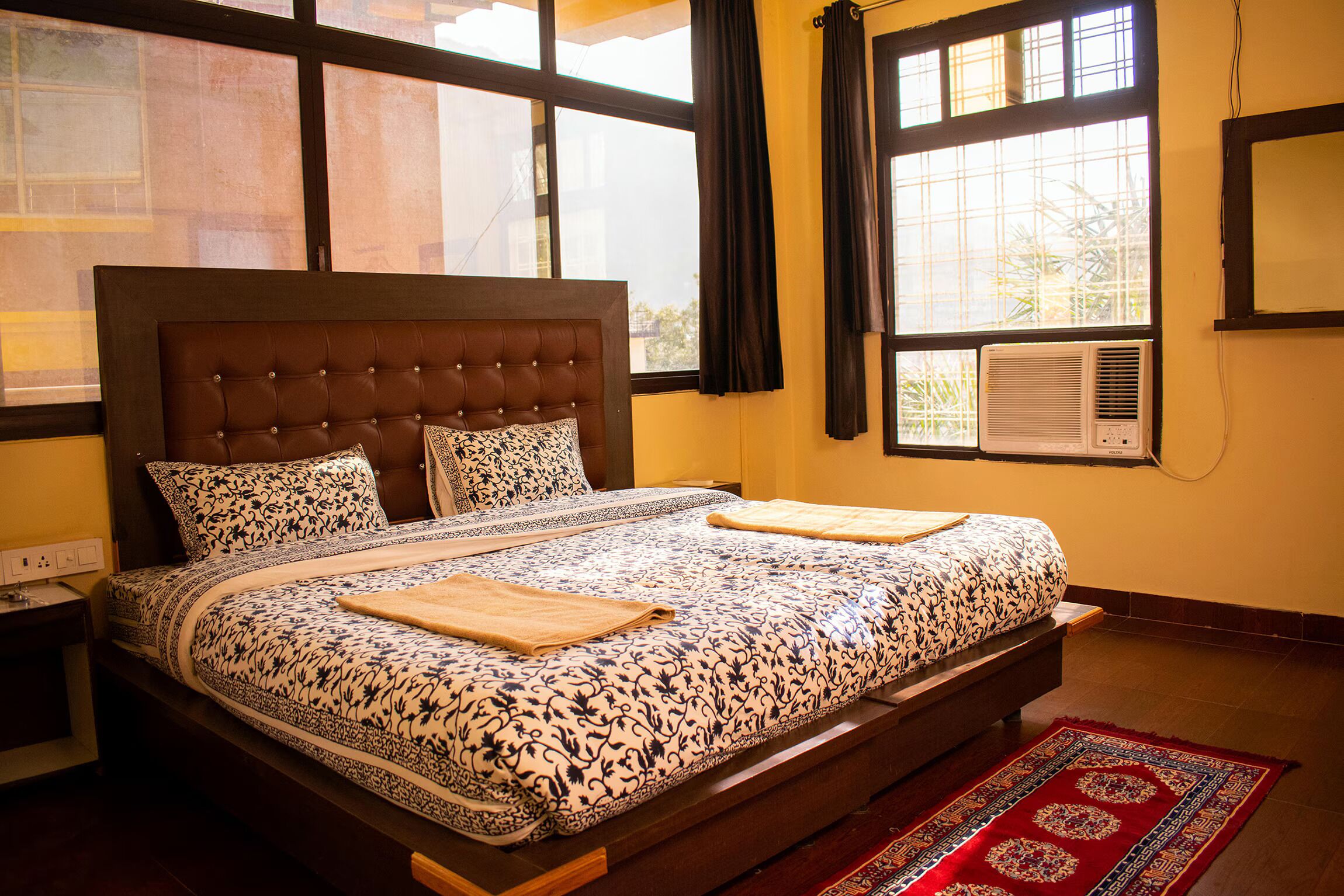 A double bed with a patterned bedspread, a wooden headboard, and a red rug.
