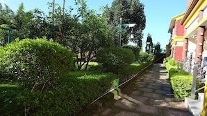 A stone pathway flanked by well-maintained hedges and trees, leading to a building with a red facade and yellow trim. - MVT Resort & Restaurant (Rishikesh)