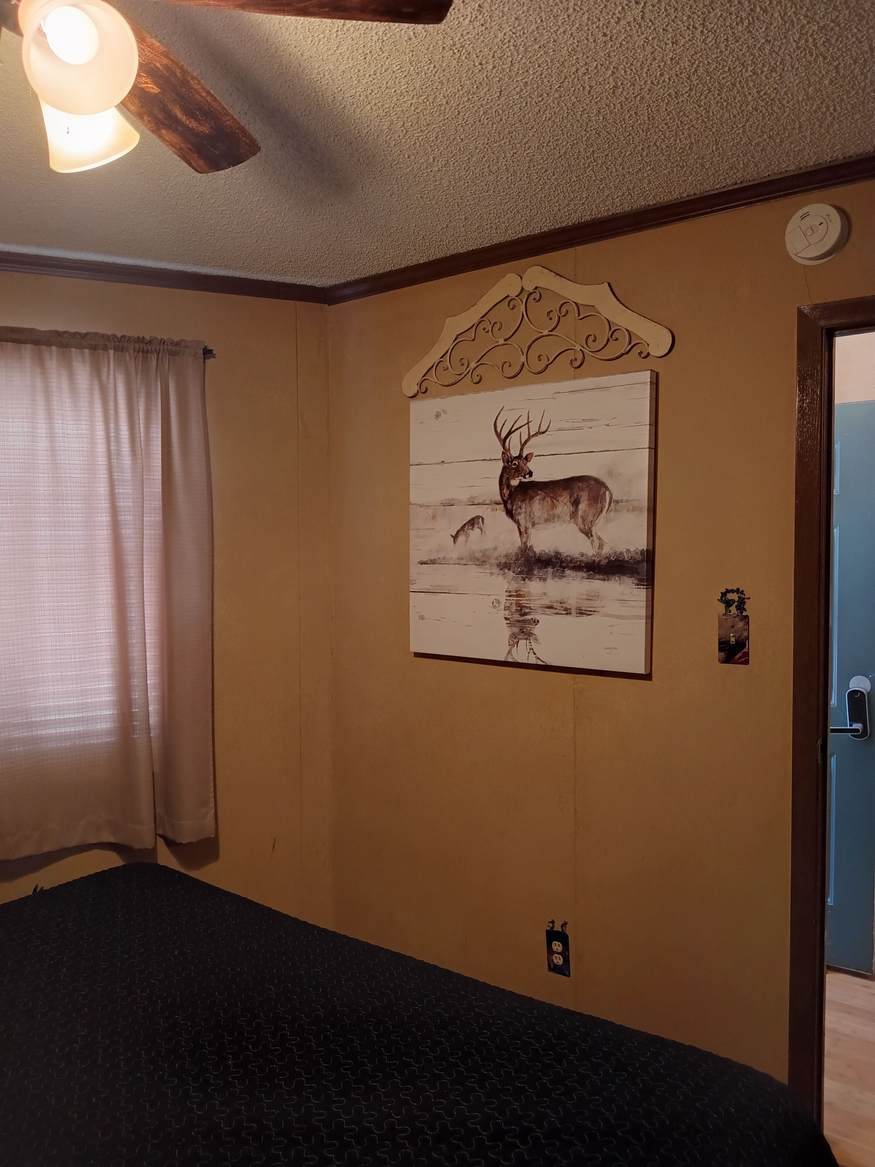 Ein Zimmer mit Deckenventilator, einem Hirschgemälde, einer Tür mit Lichtschalter und einem fenster mit Vorhängen.