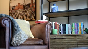 A brown leather armchair with a throw blanket, a bookshelf with books and a decorative vase, and a painting of a cityscape on the wall. - Jewel in the Garden of England - Two bedrooms (Lyminge)