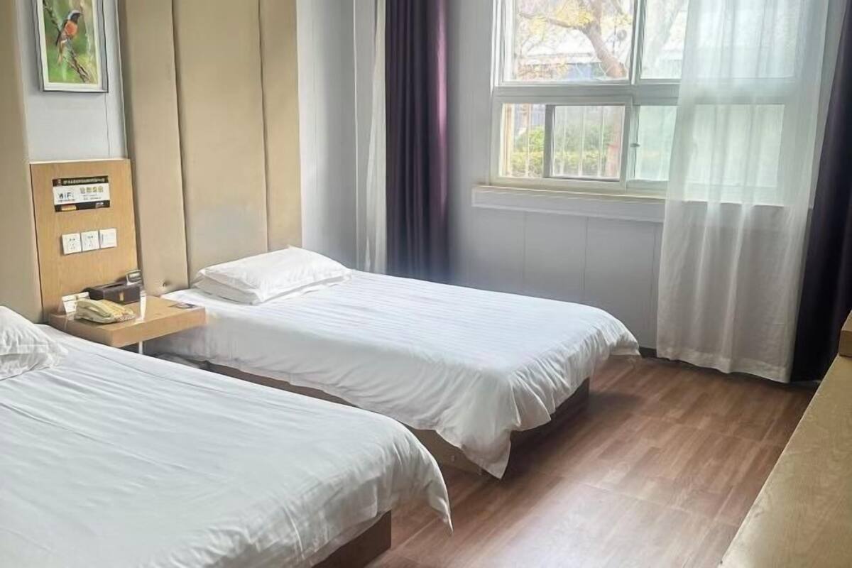 A hotel room with two single beds, a wooden nightstand, a window with curtains, and a wall-mounted panel with a sign.