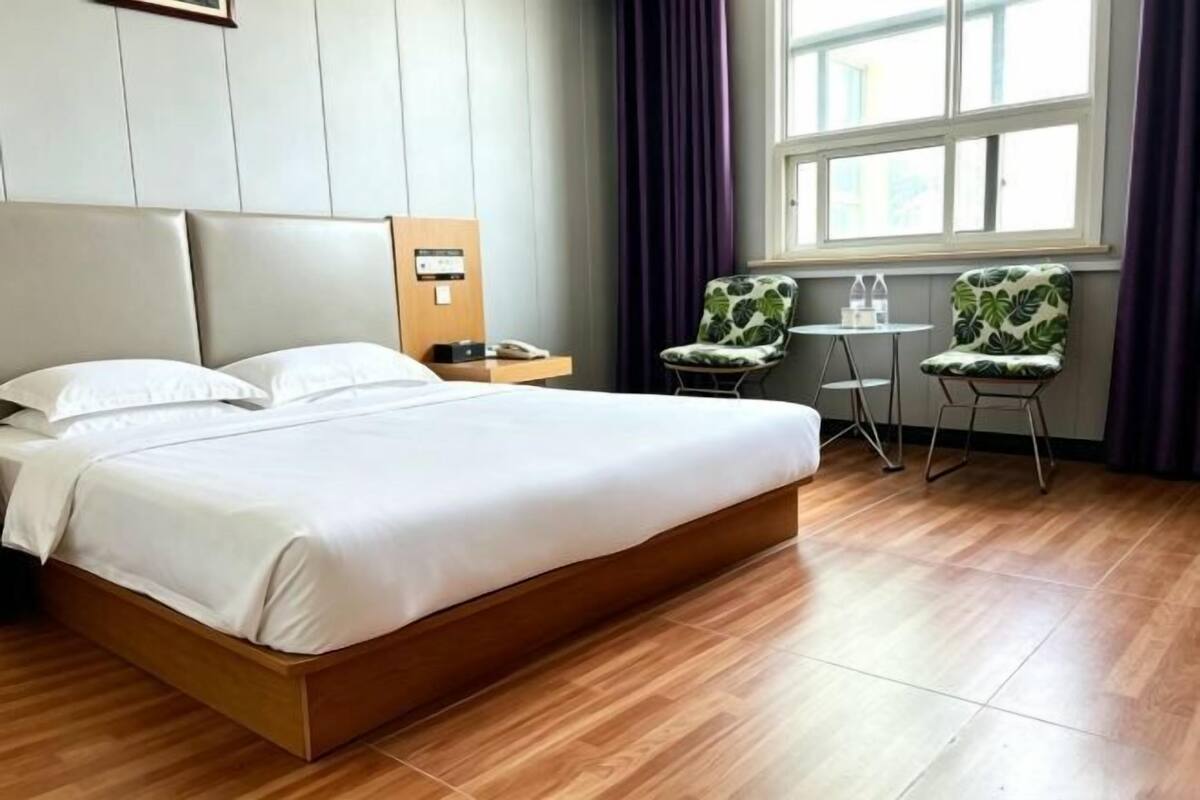 A hotel room with a bed, a bedside table, a chair, a small table, and a window with curtains.