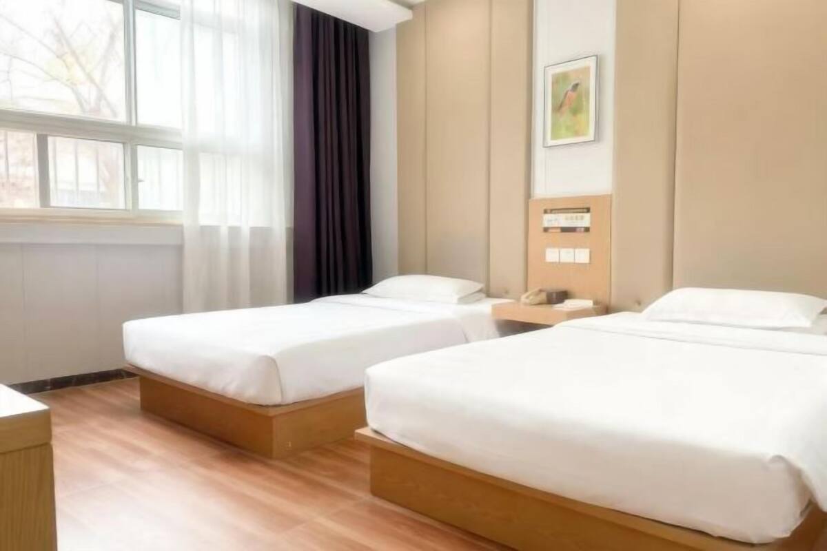 A hotel room with two beds, a wooden floor, a window with curtains, a wall-mounted phone, and a framed picture.