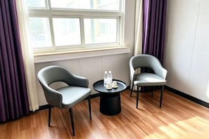 Um quarto com duas cadeiras cinzentas, uma mesinha preta com garrafas de água e uma janela com cortinas roxas.