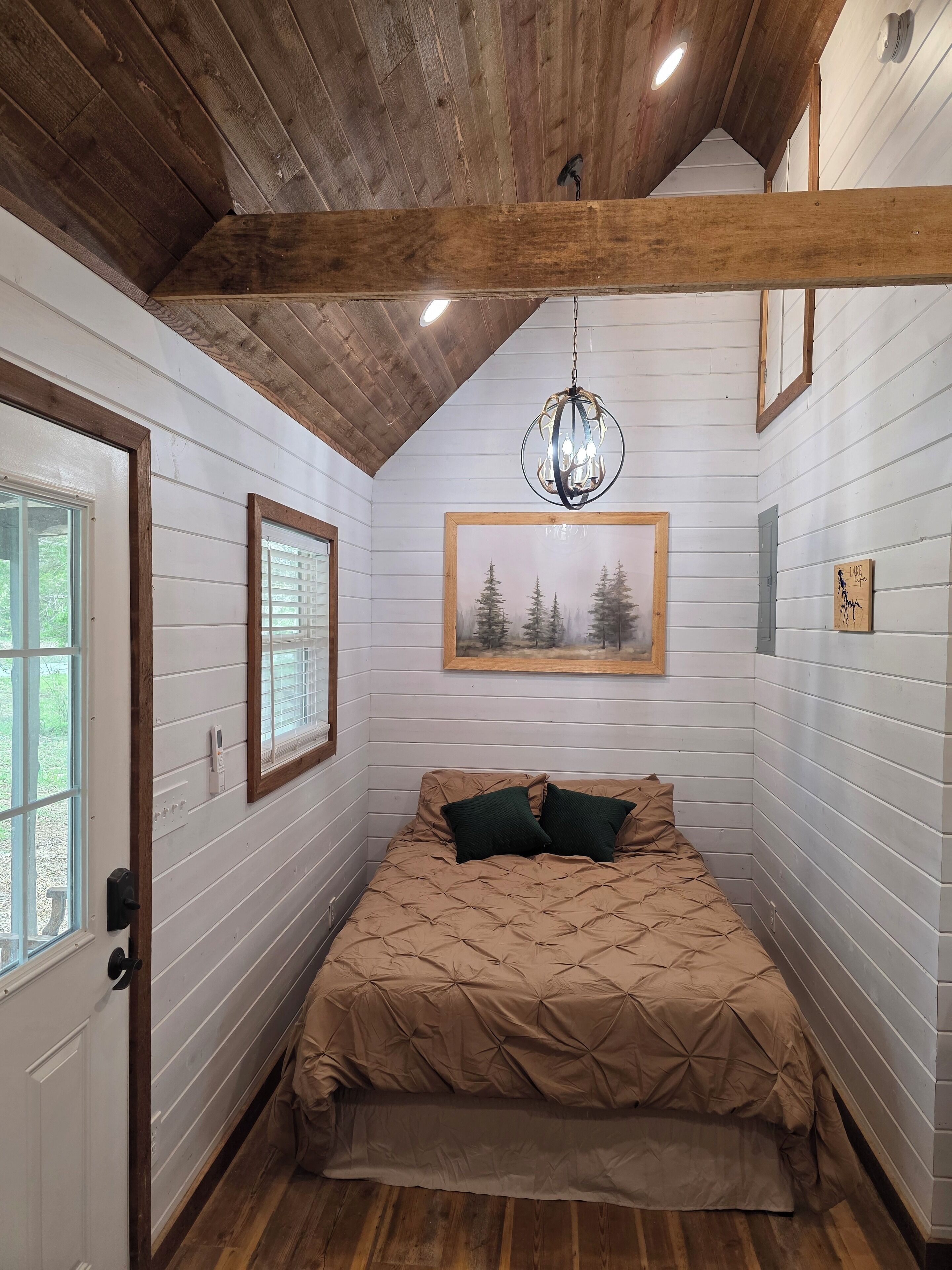 A bedroom with a wooden ceiling, a bed with a beige quilt, a chandelier, and a framed picture of trees.