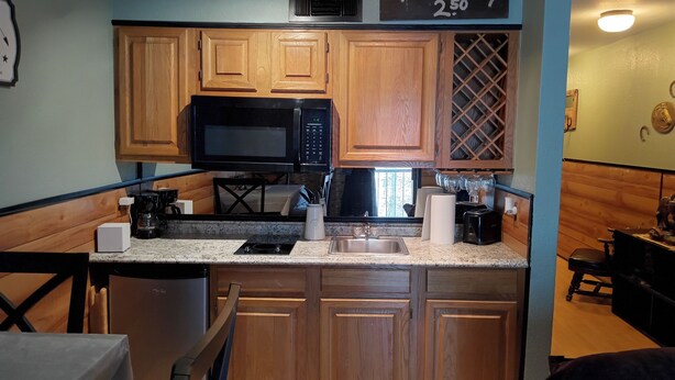 A compact kitchen with wooden cabinets, a black microwave, a sink, and a countertop with various items.