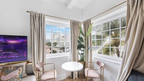 Un salon moderne avec un téléviseur, deux fauteuils roses, une table ronde et une grande fenêtre avec des rideaux.