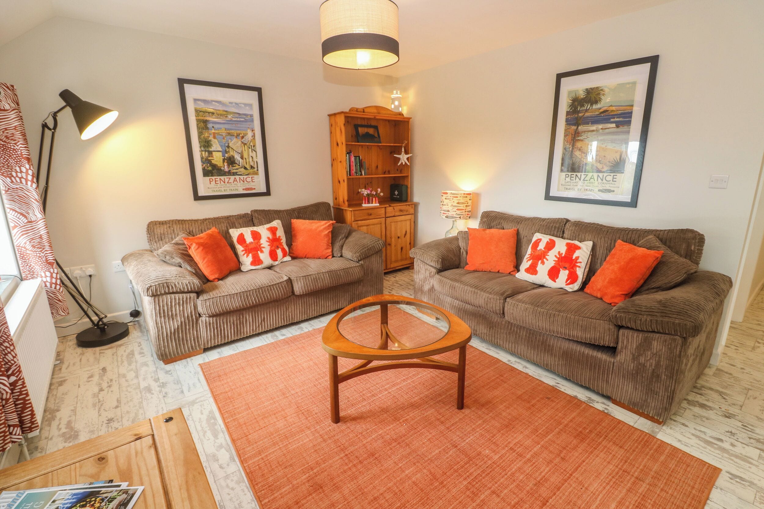 A living room with two brown sofas, orange cushions, a wooden coffee table, and framed travel posters on the wall.