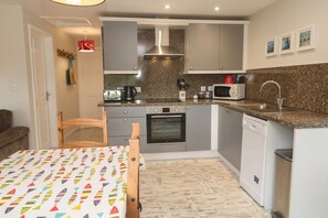 A compact kitchen with a microwave, oven, and sink. There is a dining table with a colorful tablecloth. - Sea View Loft (Penzance)