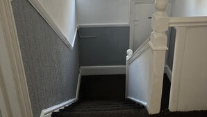 Una escalera estrecha con un pequeño ventanal en la parte superior, paredes blancas y un suelo oscuro cubierto de alfombra.