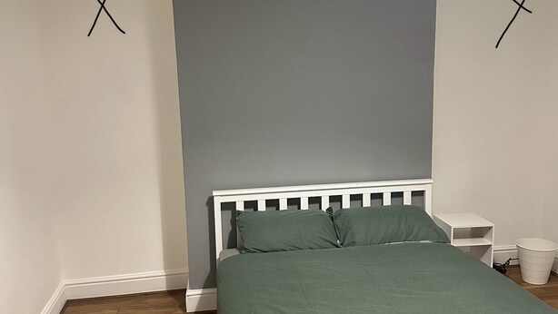Un dormitorio con estructura de cama blanca, ropa de cama verde, suelo de madera, mesita de noche blanca y un cubo blanco.