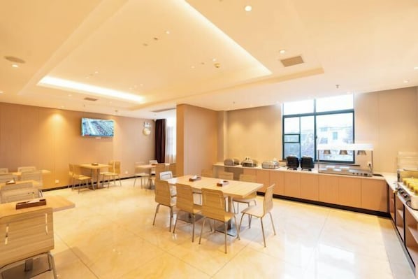 A modern hotel dining area with multiple tables and chairs, a kitchen counter, and a television mounted on the wall.