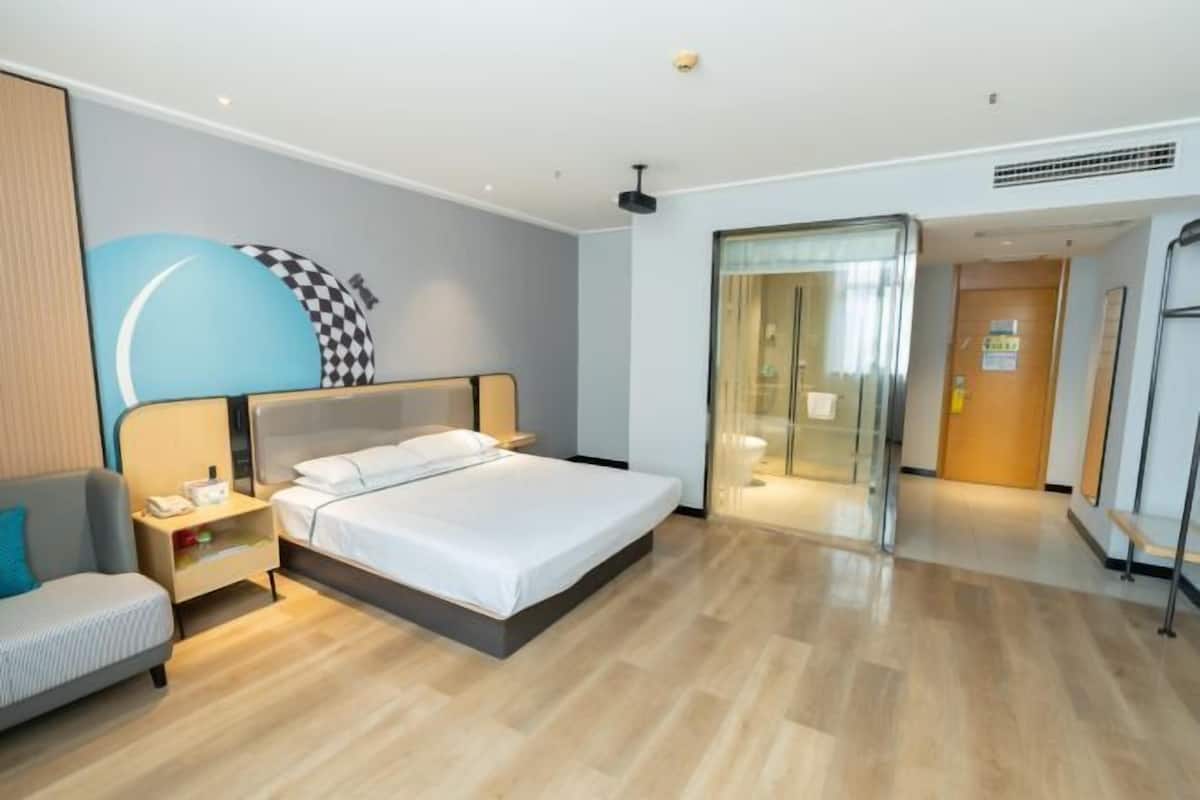 A modern hotel room with a bed, bedside table, chair, and a glass door leading to a bathroom.