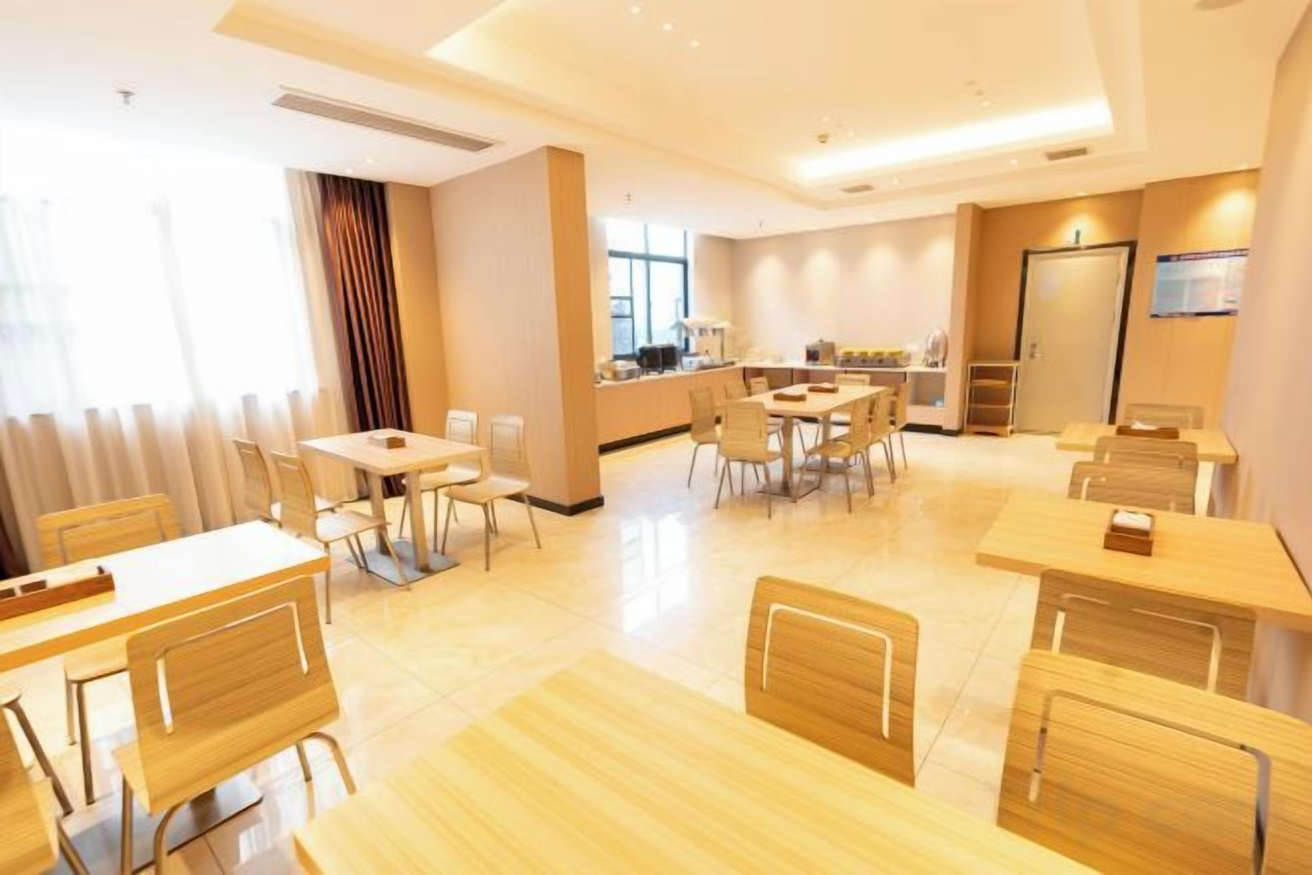 A hotel dining area with multiple tables and chairs, a kitchen in the background, and a large window providing natural light.