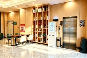 Un vestíbulo de hotel con mostrador de recepción, zona de estar, una amplia estantería, un ascensor y un cubo de basura.