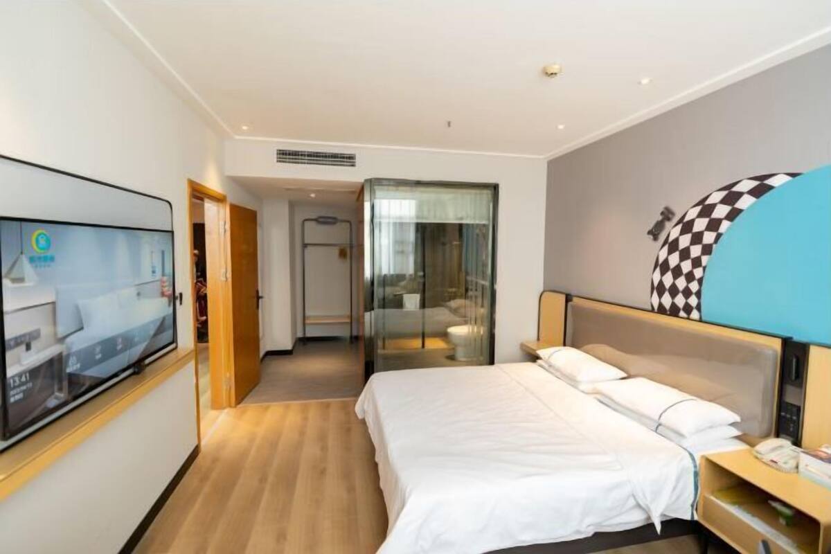 A hotel room with a bed, a TV, a desk, and a bathroom visible through a glass door.