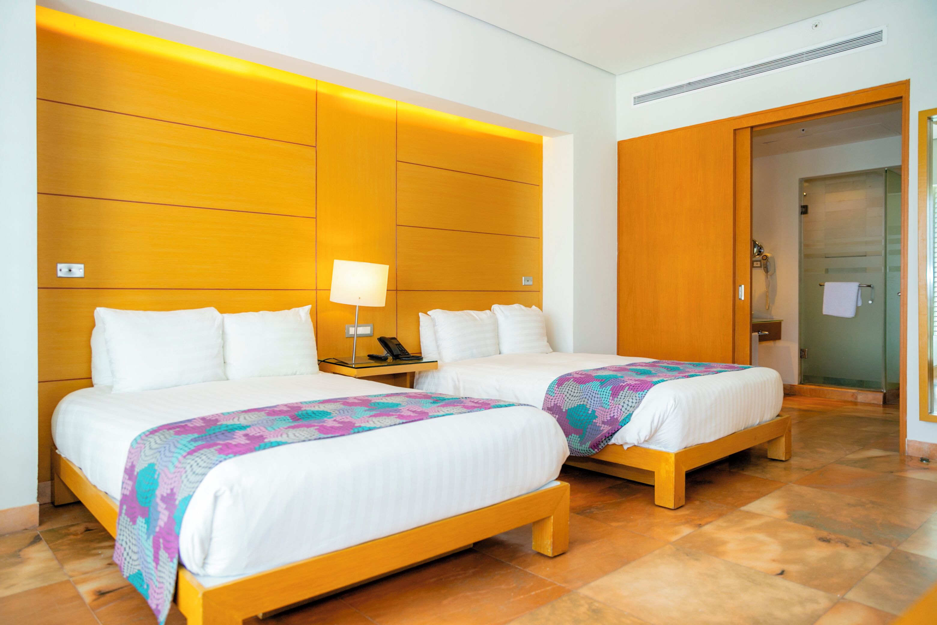 A hotel room with two beds, wooden headboards, a bedside table with a lamp, and a bathroom door.