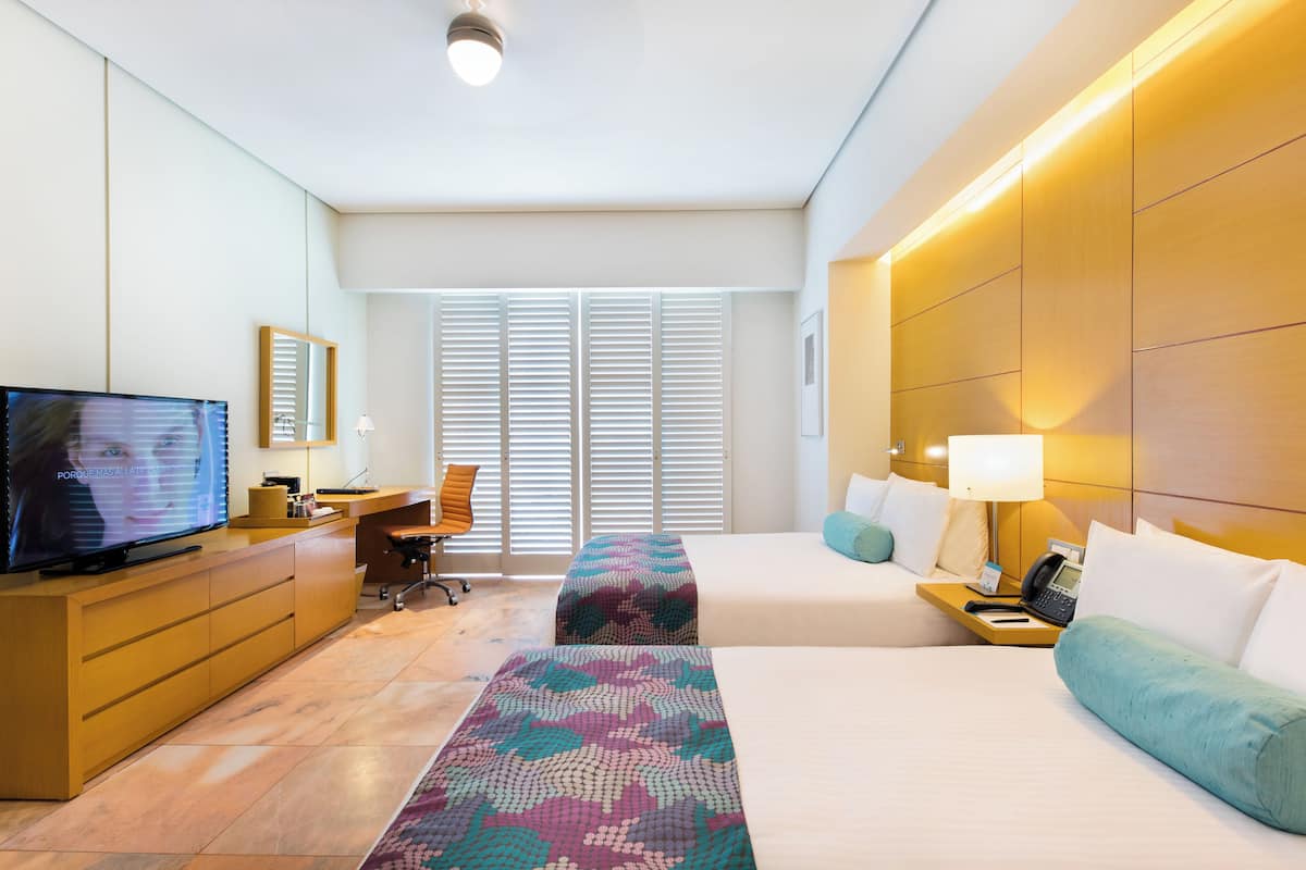 Quarto de hotel com televisão, secretária, cadeira, duas camas, mesas de cabeceira, luminárias e uma janela grande com persianas.