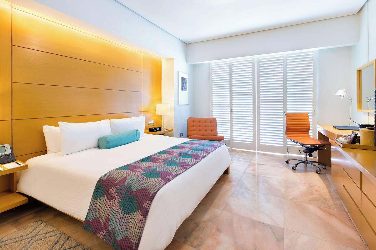 Quarto de hotel com cama, escrivaninha, cadeira, televisão e telefone.
