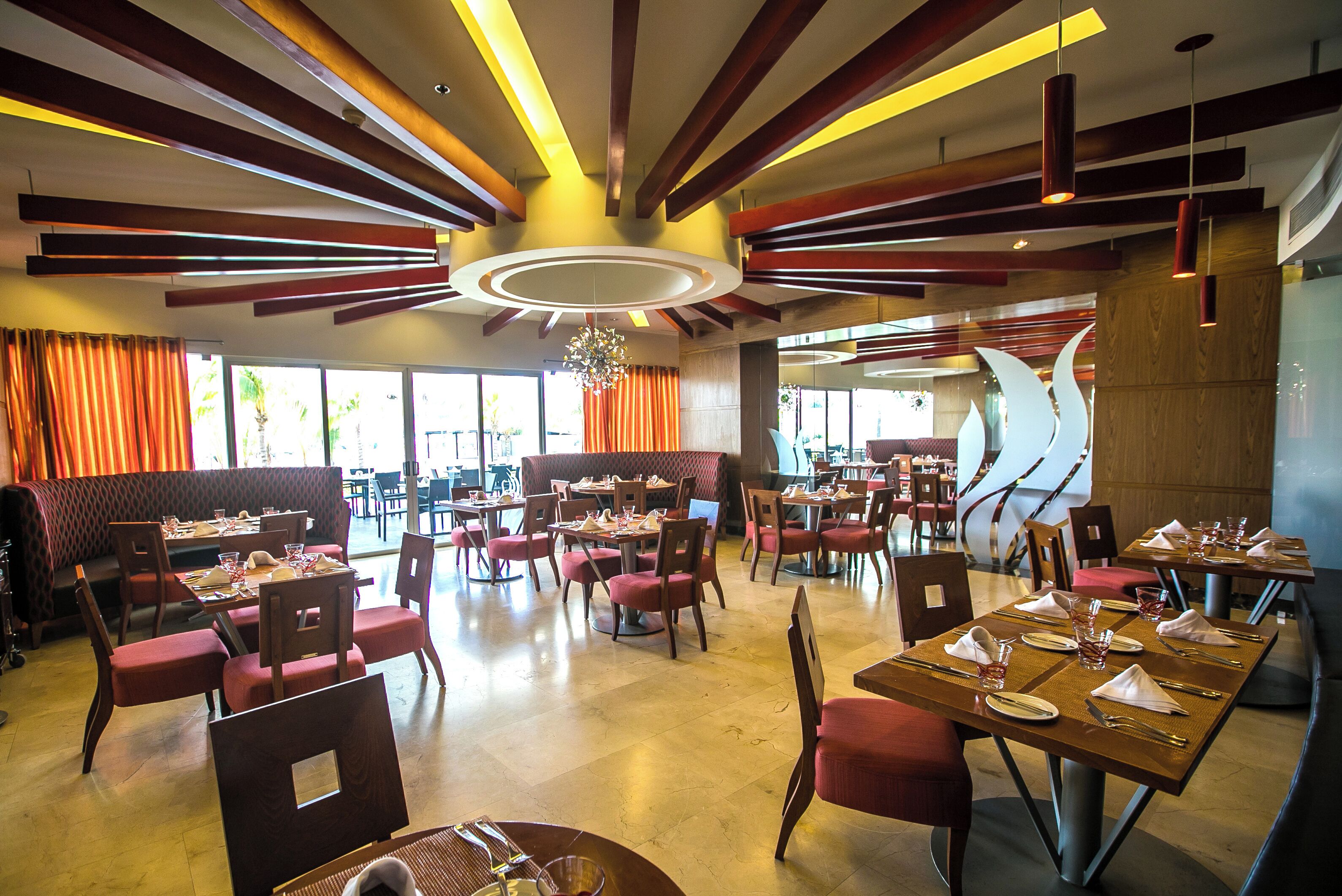 Un restaurante espacioso con varias mesas y sillas, un candelabro central y grandes ventanales que dejan entrar luz natural.
