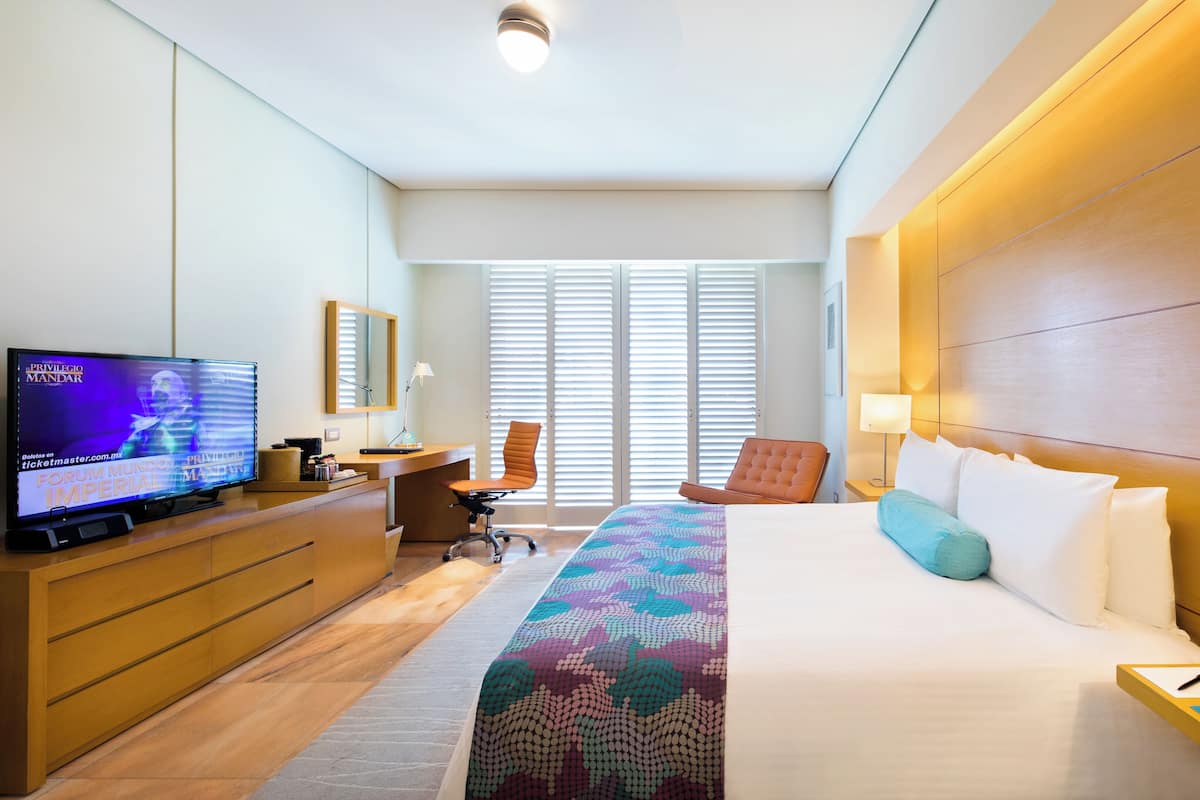 Quarto de hotel com uma cama, uma escrivaninha com televisão, uma cadeira, uma mesa de cabeceira com telefone e uma janela com persianas.