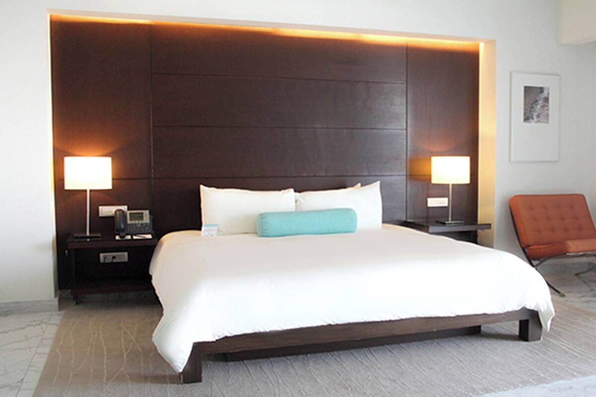Uma cama grande com cabeceira escura, duas mesas de cabeceira com luminárias, uma poltrona laranja e um quadro na parede.