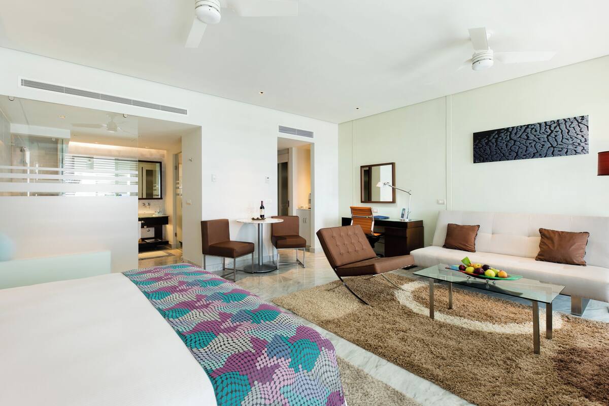 Quarto de hotel moderno com cama, sofá, mesa de centro e área de refeições.