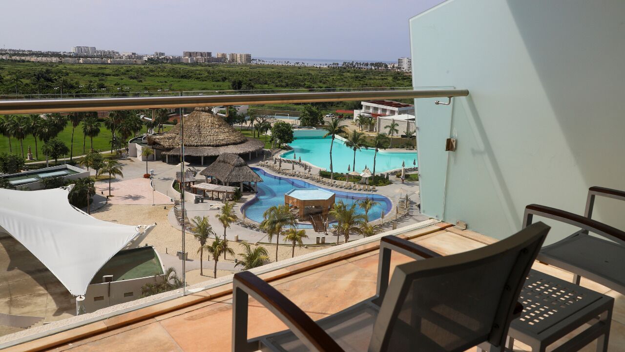 Un balcón con vista al área de la piscina de un resort, incluyendo una cabaña con techo de paja y sillas de descanso.