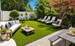 Ein Garten mit Rasen, einem weißen Zaun, einem Holztisch, einer Couch und Liegestühlen.