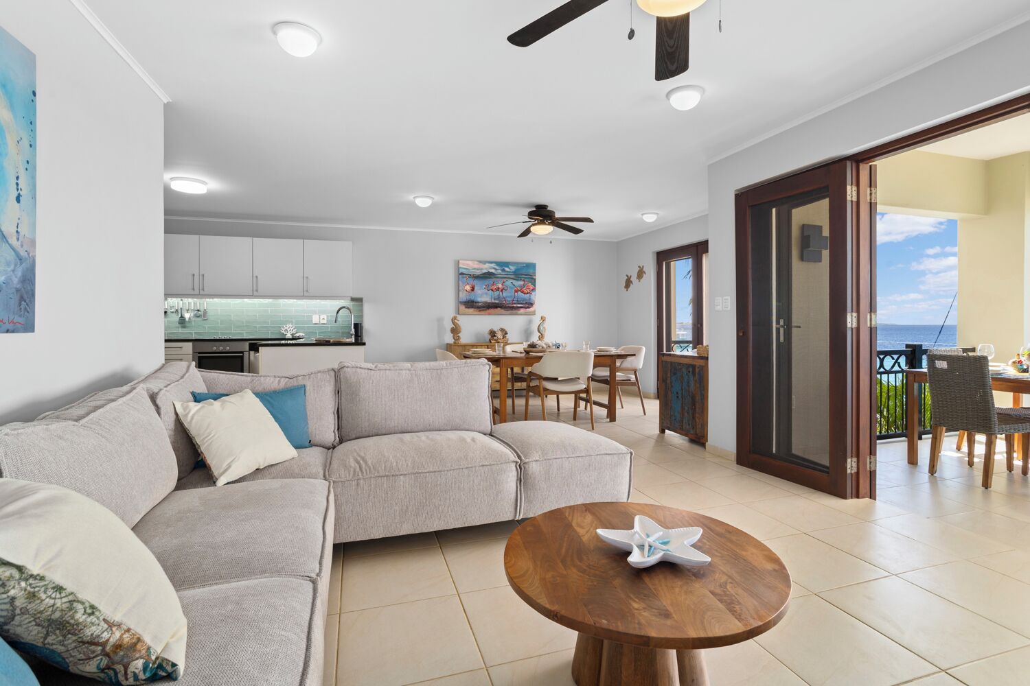 A modern living room with a sofa, a wooden coffee table, and a dining area with a table and chairs. There is a kitchen in the background with white cabinets and a tiled backsplash.