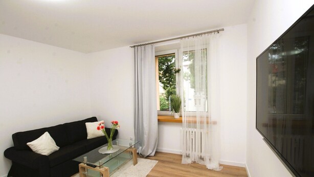 Uma sala de estar com um sofá preto, uma mesa de centro de vidro, um tapete branco, uma TV na parede e uma janela com cortinas leves.
