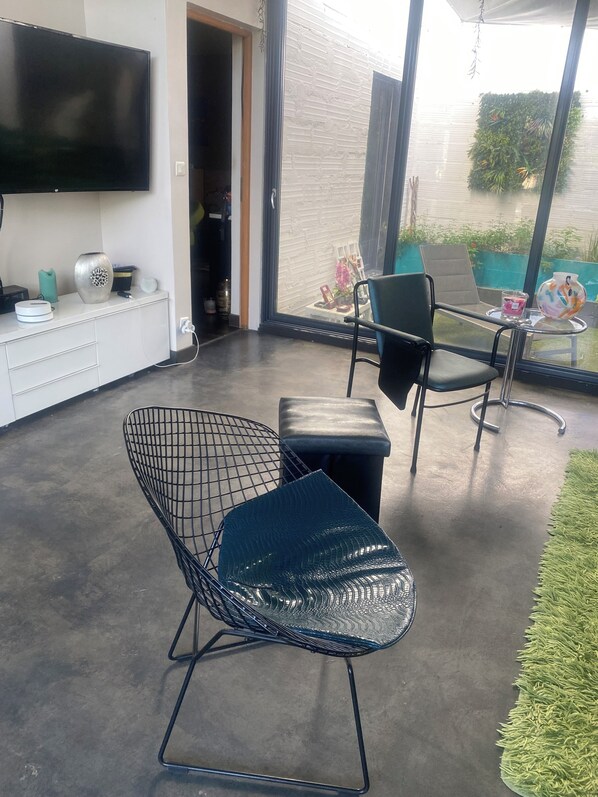 A modern outdoor seating area with a metal chair and a glass table, a flat-screen TV mounted on a white cabinet, and a view of a garden through large windows.
