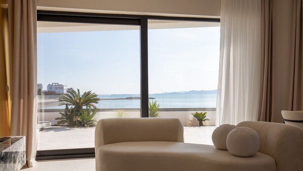 A modern living room with a large window offering a view of the sea and distant buildings. - Villa Noles (Sukošan)
