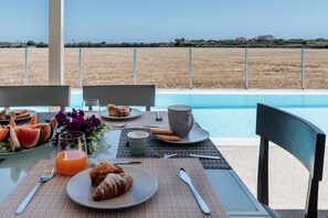 Une table de petit-déjeuner avec des croissants, des tranches de pastèque et du jus d’orange, donnant sur une piscine et un champ.