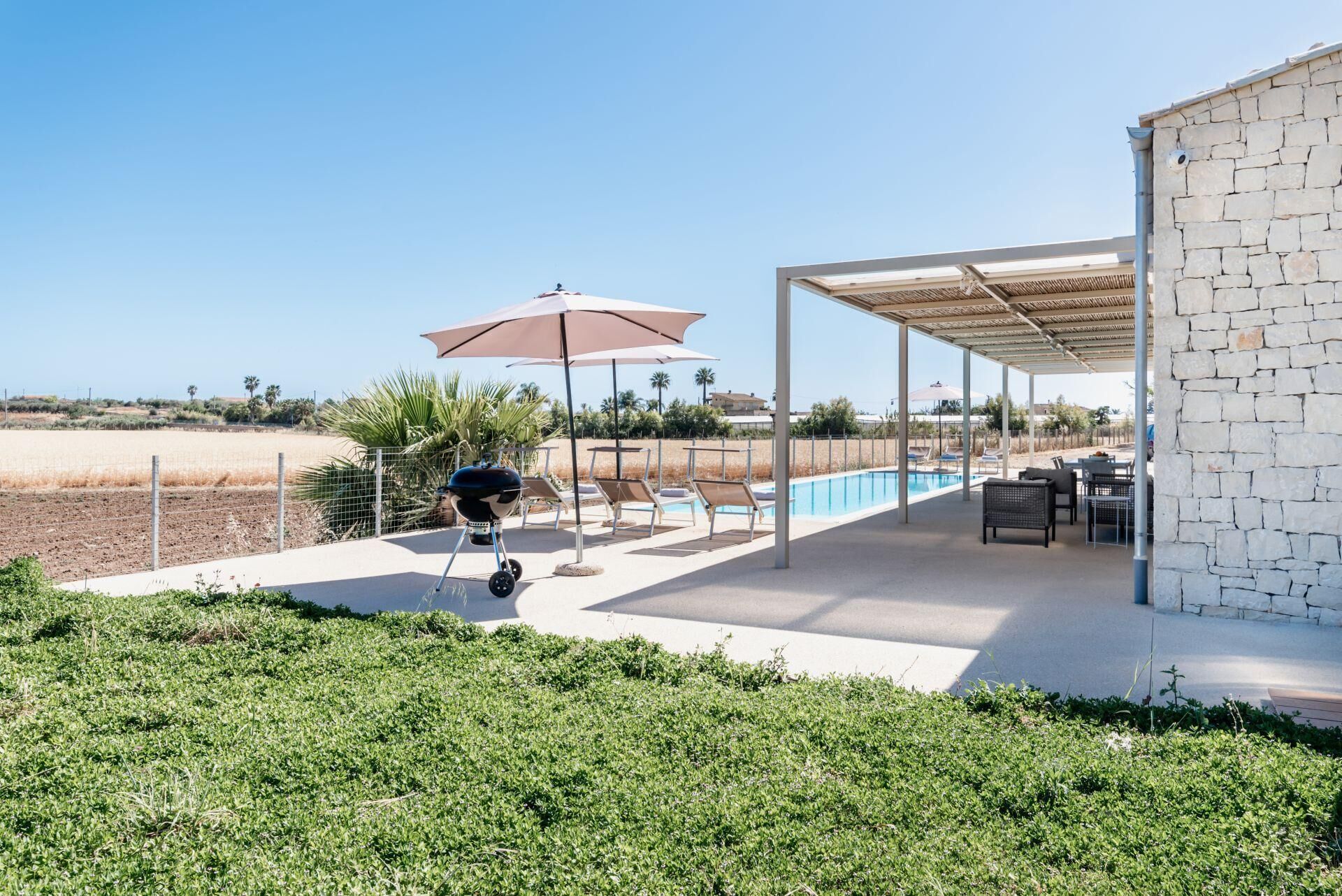 A covered outdoor area with a barbecue, lounge chairs, and a pool.