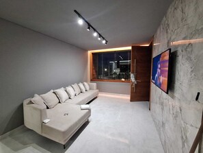 Un salon moderne avec un canapé, un téléviseur fixé au mur et une fenêtre donnant sur la ville illuminée la nuit.
