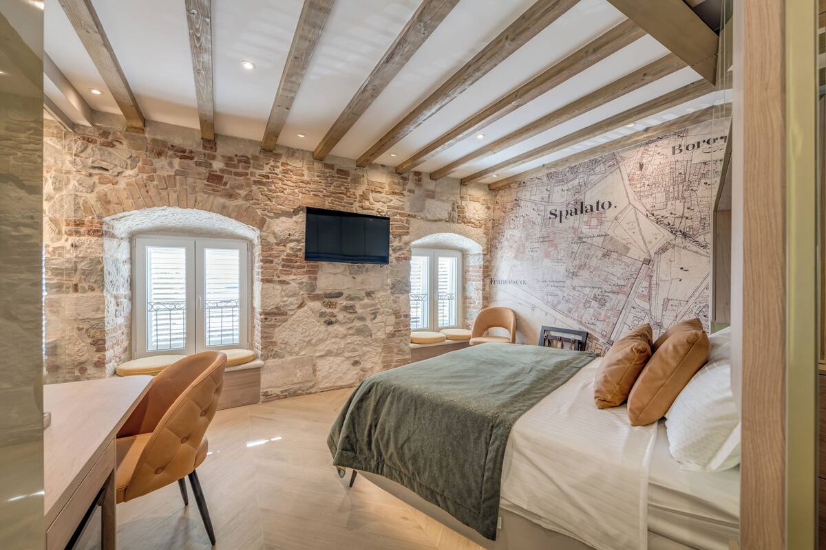 A bedroom with stone walls, a bed with pillows, a desk, and a TV mounted on the wall.