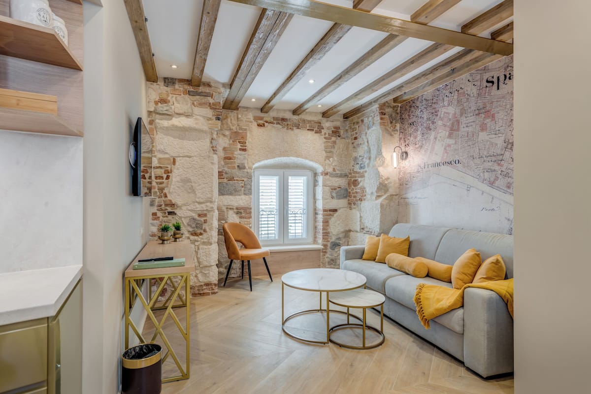 A cozy living room with exposed wooden beams, a stone wall, a map mural, a grey sofa with yellow cushions, and a round coffee table.