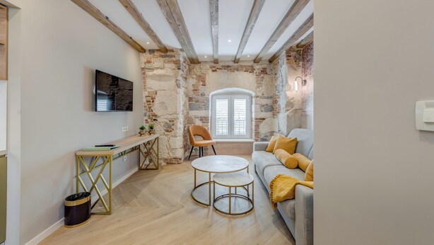 Ein gemütliches Wohnzimmer mit freiliegenden Holzbalken, einer Steinwand, einer Couch mit gelben Kissen, einem runden Couchtisch und einem an der Wand montierten Fernseher.
