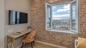 Ein Zimmer mit Backsteinwand, einem Schreibtisch mit Stuhl, einem an der Wand befestigten Fernseher und einem Fenster mit Meerblick.