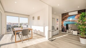 A modern living room with a TV, a dining area with a table and chairs, and a balcony with a view. - GuestReady - Quinta do Moledo House (Charneca de Caparica)