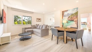 A modern living room with a sofa, a dining table, and chairs. There is a painting on the wall and a window with a view of trees. - GuestReady - Quinta do Moledo House (Charneca de Caparica)