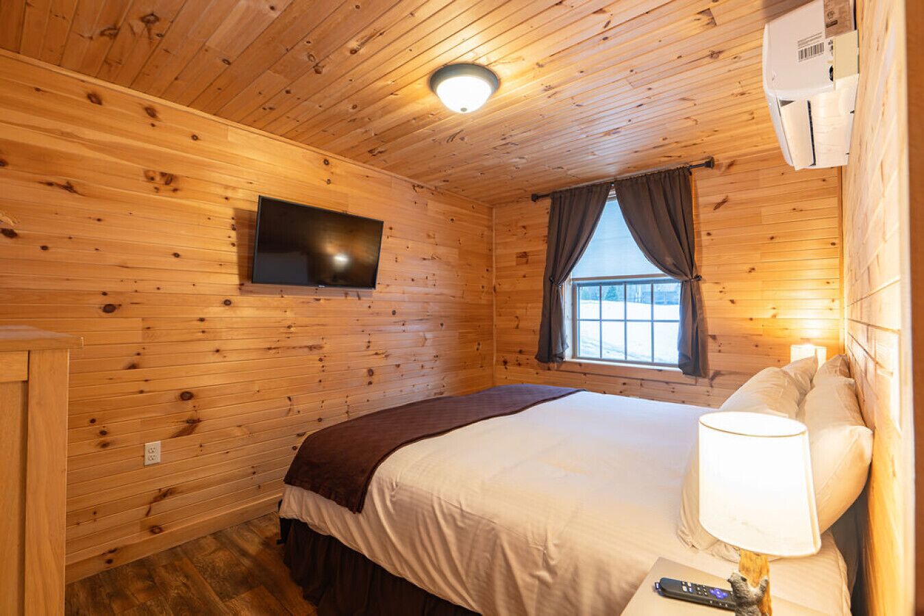 A wooden-paneled room with a bed, a TV mounted on the wall, and a window with curtains.