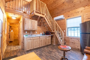 A cabin interior with a wooden staircase, kitchen with wooden cabinets, a dining table, and a refrigerator.
