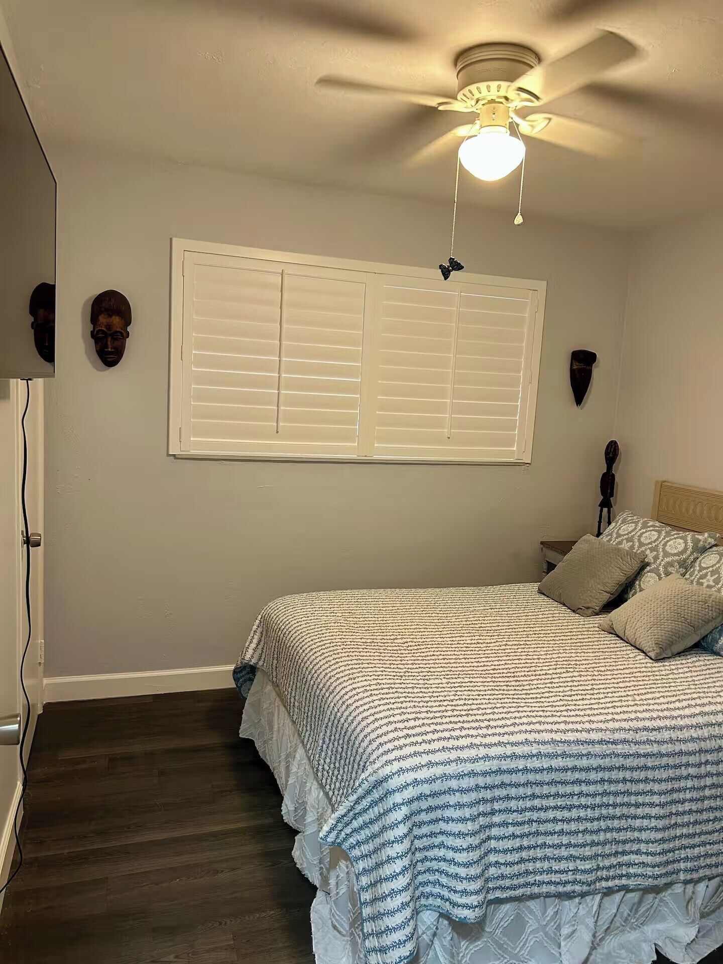 Une chambre à coucher comprenant un lit, un ventilateur de plafond, une fenêtre avec des stores et un luminaire fixé au mur.
